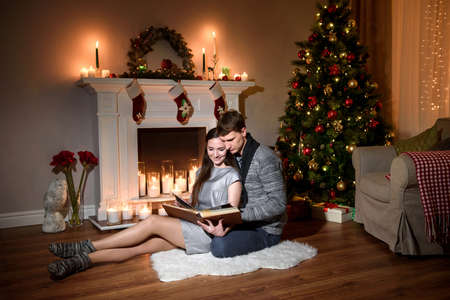 A young family looking through a family photo album in a cozy new year living room. Nice love couple sitting on carpet in front of fireplace. Woman and man celebrating Christmas.の写真素材