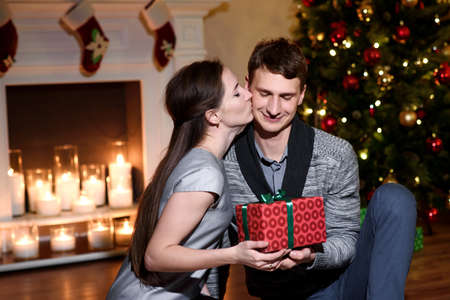 Young couple with gifts in front of Christmas tree. A young wife is giving her husband a new year present and kissing his cheek, he is cutely embarrassed.の写真素材