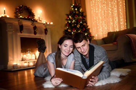 A young family spending evening together looking through some pictures in a big photo album. Couple hugging in front of fireplace at home, reading book.の写真素材
