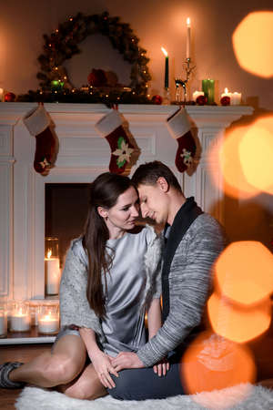 A lovely couple cuddling gently on a white furry carpet near a christmas decorated fireplace, sunny bunnies playing on a picture. Young couple sitting on floor at burning fireplace.の写真素材