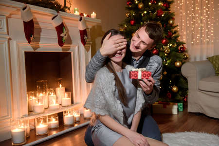 A young husband is making a surprise present to his beloved, romantic christmas. Merry Christmas. Young couple celebrating Christmas at home. Cozy moments in winter holidays at decorated tree.の写真素材