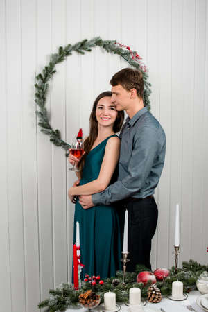 A young husband hugging his elegantly dressed wife from her back, she is smiling happily holding a glass. Beautiful young couple is holding glasses of wine and smiling while celebrating at home.の写真素材