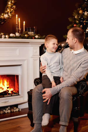 Father with his son sitting in armchair by Christmas tree and fireplace, looking at each other. Christmas wreath on the wall. On the fireplace there are candles and books, Xmas tree decorated by lightsの写真素材