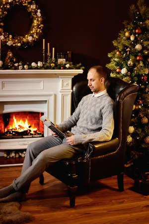Caucasian man in gray clothes sits in chair by burning fireplace and reads magazine. Warm and cozy evening in Christmas room interior design, Xmas tree decorated by lights presents gifts toys, candlesの写真素材