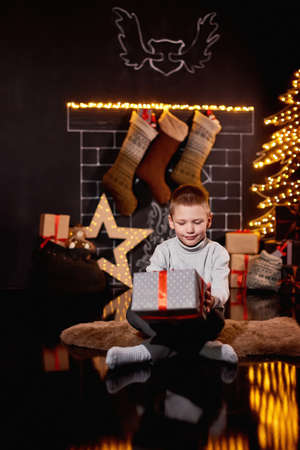 Merry Christmas! Happy kid with magic gift at home near Christmas tree and fireplace. A bag with gifts, a star, socks hanging on the fireplace. Xmas winter holiday concept. Funny kid playing at homeの写真素材