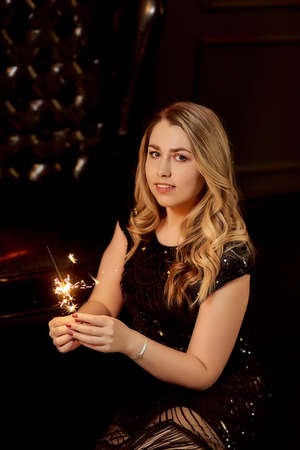 Beautiful slender girl blonde in a black dress holds a Bengali fire, looking at the frame, sitting on a black leather chair. The concept of holiday, Christmas, party, vertical photo, place for textの写真素材