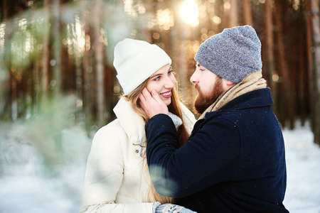 Smiling girl with long hair in a white puff jacket, knitted cap and bearded guy in a black winter coat hugs each other in a snowy pine forest outdoor. Romantic winter holiday concept. Free copy spaceの写真素材