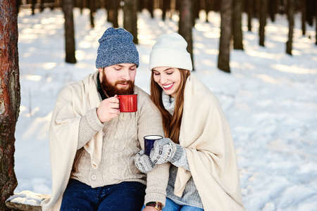 Close up side profile photo of beautiful couple in winter park with hot drinks warms themselves covered by a white plaid. Winter holidays concept. Winter vacation, holiday, caucasianの写真素材