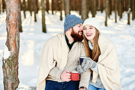Romantic relationship and winter holiday vacation concept. Happy smiling couple under warm plaid with cups of hot drinks hugging and kissing outdoors in winter forest. Romantic couple in love feelingの写真素材