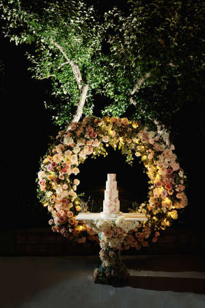 Wedding cake in the evening. Cake on the background of an arch of flowers. White and Pink colors of cream and decoration. Fourlevels of different sizes. Night wedding ceremonyの写真素材