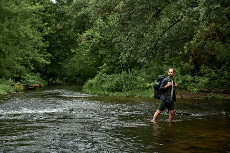 A man in a plaid shirt and green shorts with a large hiking backpack looking back at the far bank of...