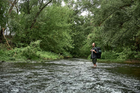 A man in a plaid shirt and green shorts with a large hiking backpack crosses the river, looking up. Equipment, gear for hiking. New routes for recreation, camping in summer, outdoor travel concept.の写真素材