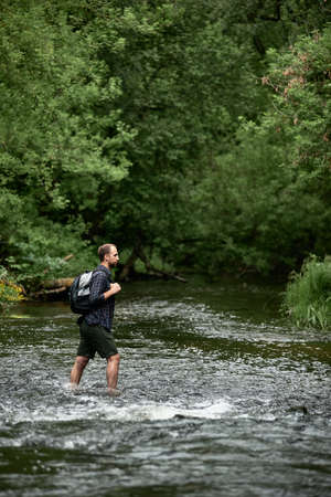 A man in a plaid shirt and green shorts with a backpack crosses the river, looking at the far bank of the river. The concept of hiking trips. Equipment, gear for hiking. Space for text, verticalの写真素材