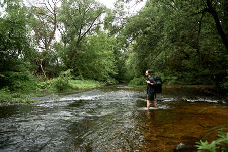 A man in a plaid shirt and green shorts with a large hiking backpack crosses the river, looking at the far bank of the river. The concept of hiking trips. Equipment, gear for hiking. Space for textの写真素材