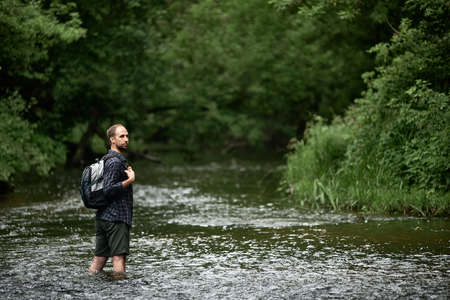 Back view of a hiker in a plaid shirt with a backpack crossing a river looking back at the far bank of the river. Equipment, gear for hiking, Wanderlust lifestyle, vacations in the wilderness.の写真素材
