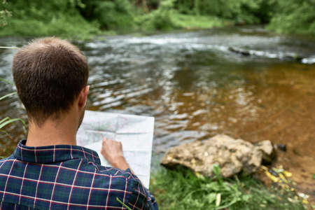 The man in a plaid shirt standing on the bank of the river points a finger to a place on the map....