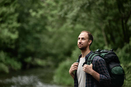 Attractive male hiker with a large backpack standing on the bank of the river and enjoying nature, looking up. The concept of hiking trips wanderlust lifestyle, vacations outdoors in the wilderness.の写真素材