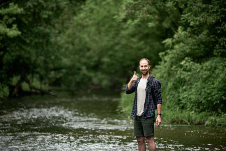 A man in a plaid shirt with a backpack crosses the river, looking and smiling at camera and shows thumbs up. The concept of hiking trips, adventure, good gear for travel. Space for textの写真素材