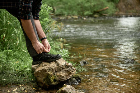 Hands of male hiker tying shoelaces placing his foot on a large stone by the forest stream, while trekking outdoors, shallow debth of field, selective focus. The concept of comfortable hiking shoes.の写真素材