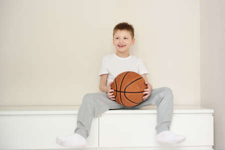 Cute smiling boy in casual clothes sitting on the dresser in his room holding a basketball. White background. Space for textの写真素材