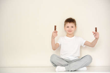 Cute smiling boy in casual clothes sitting on the dresser in his room holding a chocolate. Little boy of six years from a chocolate bar isolated on white background.の写真素材