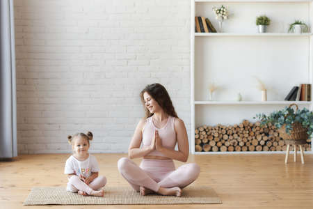 Smiling young curly woman looking with love at her small kids girl sitting on the carpet floor in lotus pose namaste hands, have fun and doing yoga exercise. Keep calm with kids, healthcare conceptの写真素材
