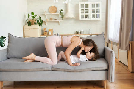 Happy young mother in light pink sportswear with curly long hair holding and kissing cute daughter lying on couch at home. Close up relaxing parent having fun free time and playing with child.の写真素材