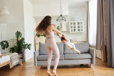 Active family fooling and spining around in cozy light living room, young mother holding hands little cheerful daughter, enjoying music dancing at home, leisure activity, weekend, free time concept.の写真素材