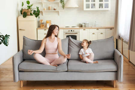 Smiling active young woman in light pink outfit lookng at her little daughter sit on couch practicing yoga in lotus position meditat? with mudra hands at living room, happy funny harmony relatonship.の写真素材