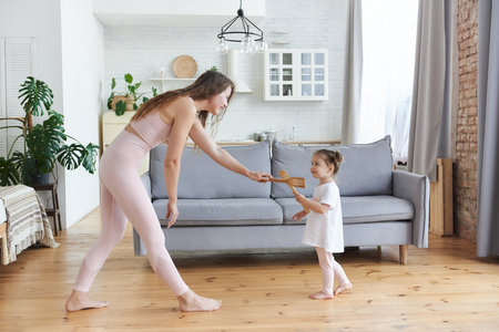 Excited mother and little daughter dancing and playing with using kitchen appliances in cozy light living room at home, family having fun moving barefoot with on the kitchen.Free time together conceptの写真素材