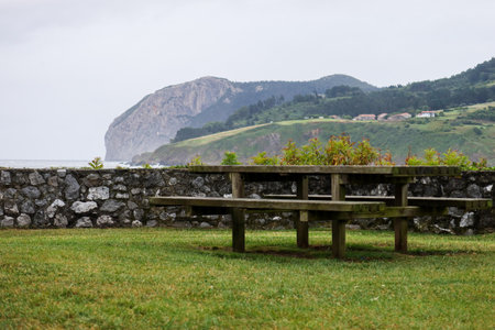 A tranquil scene in Mundaka shows a green park with a stone wall and a small table. Beyond the wall, the vast ocean stretches out, framed by distant green hills under a cloudy sky.の写真素材