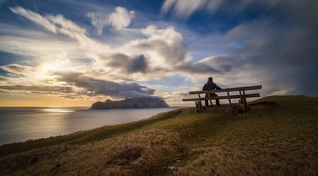 Man on bench at Gasadalur village and Mykines Island in sunset, Faroe Islandsの写真素材
