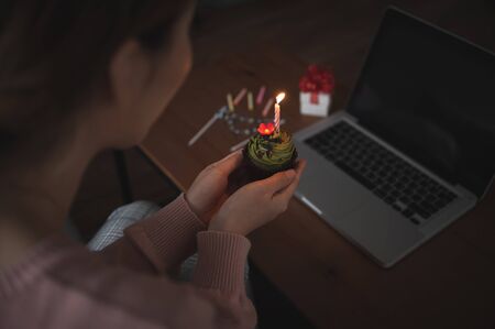 Asian woman celebrating colleague birthday through video call online virtual party at home while isolated stay at home. hand holding cup cake light up candleの写真素材