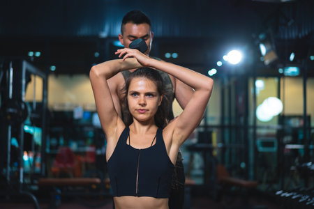 Caucasian young woman in sportswear lifting dumbbell  with trainer care for safety in fitness club...