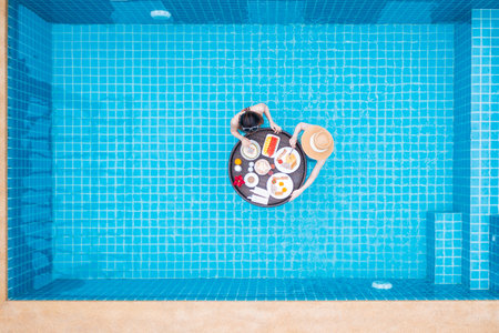 Top view of two women eating breakfast on floating tray in swimming pool in the luxury villa resort. Shot by drone.の写真素材