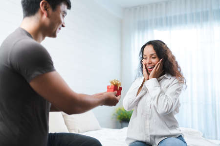 Happy asian man giving gift box to his girlfriend while sitting on bed at homeの写真素材