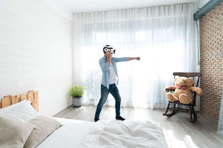 Man in VR glasses dancing on the bed at home. Concept of modern technology and entertainment.の写真素材