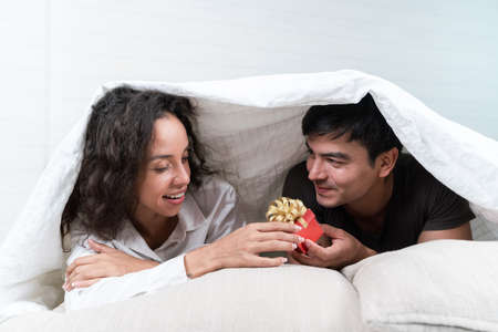 Happy young couple in love lying under blanket and holding a gift boxの写真素材
