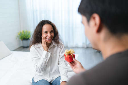 Smiling woman holding gift box while sitting on bed with her boyfriendの写真素材