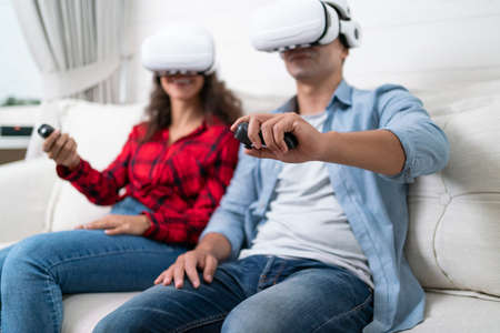 Young couple playing video games with virtual reality headset and controller sitting on sofa at homeの写真素材