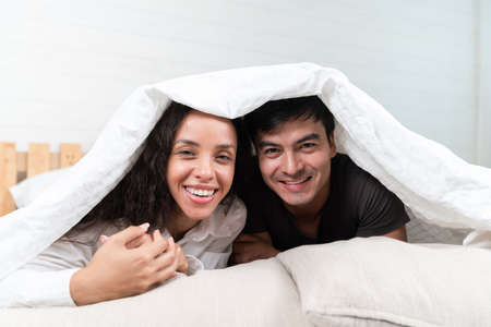 Cheerful Hispanic Couple Relaxing Under Blanket In Bedの写真素材