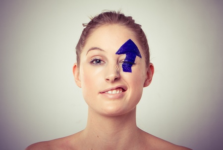 Portrait of smiling young blond haired woman with upward pointing arrow painted over eye, white...