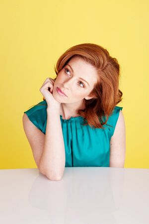 Beautiful redhead woman seated at a table with her chin on her hand lost in deep thought.の写真素材