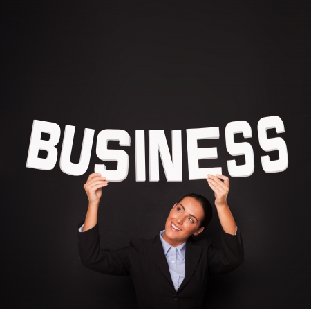 Smiling beautiful professional businesswoman holding up the word BUSINESS in white lettering on a black studio backgroundの写真素材