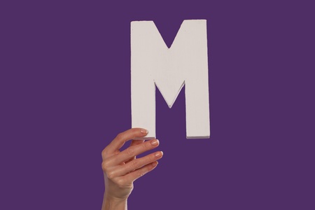 Female hand holding up the uppercase capital letter M isolated against a purple background conceptual of the alphabet, writing, literature and typefaceの写真素材