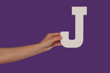 Female hand holding up the uppercase capital letter J isolated against a purple background conceptual of the alphabet, writing, literature and typefaceの写真素材