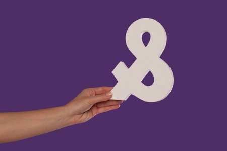 Female hand holding up a white ampersand symbol isolated against a purple background signifying plus, and, in conjunction with, or jointlyの写真素材