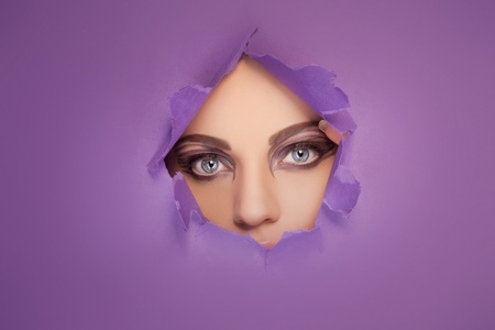 Woman with beautiful eyes and creative eye makeup looking through a hole in a sheet of purple paper so that only her eyes are visibleの写真素材