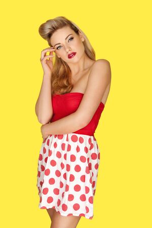 Glamorous blonde retro fashion model in a red polka dot miniskirt with vivid red lipstick and an olden day coiffure posing against a yellow studio backgroundの写真素材