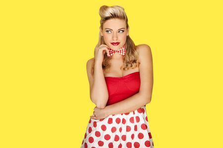Glamorous blonde retro fashion model in a red polka dot miniskirt with vivid red lipstick and an olden day coiffure posing against a yellow studio backgroundの写真素材